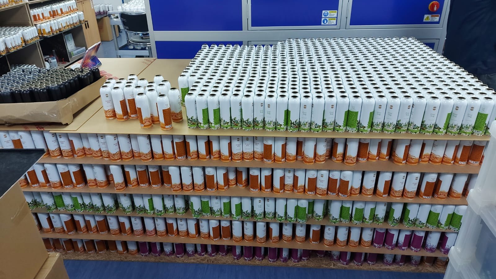 Printed Ajmal body spray cans stacked in layers at the studio, the Amber Tide, Pear Hug and Mystic Bliss designs side by side from the same production run