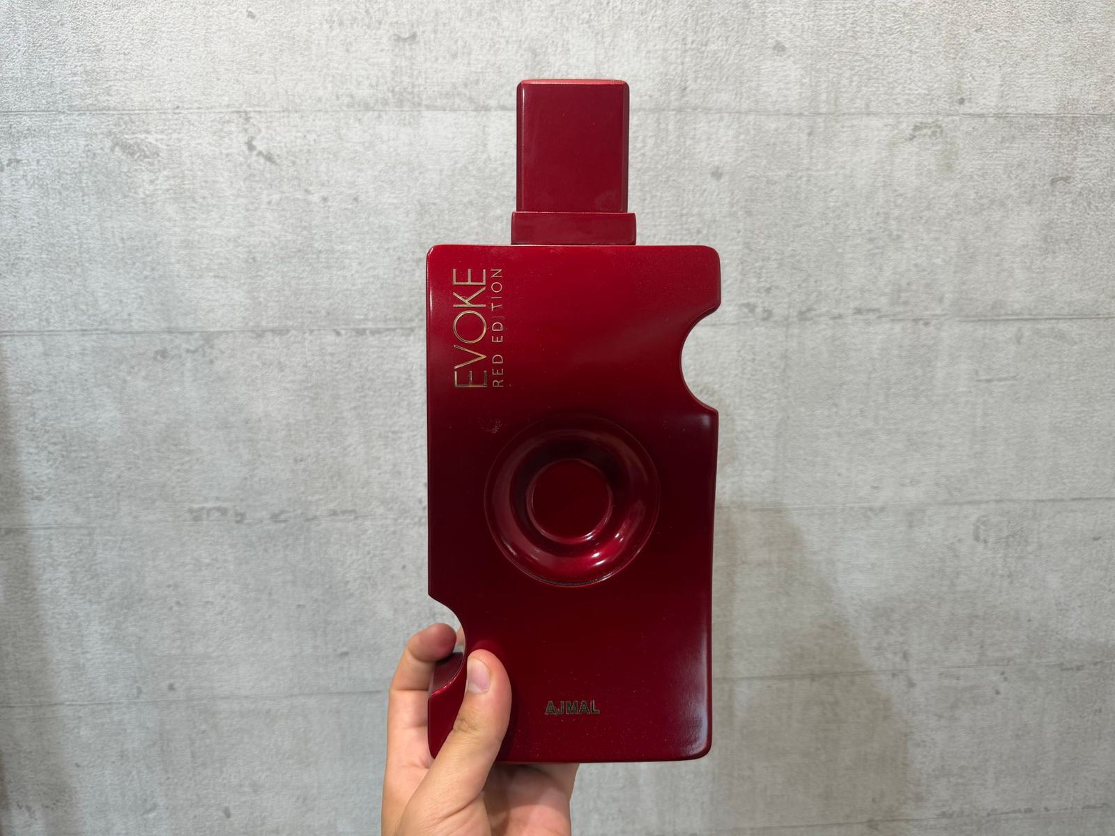 مجسم عملاق لزجاجة عطر Ajmal Evoke Red Edition تحمله يد واحدة أمام جدار خرساني، ما يُظهر حجمه