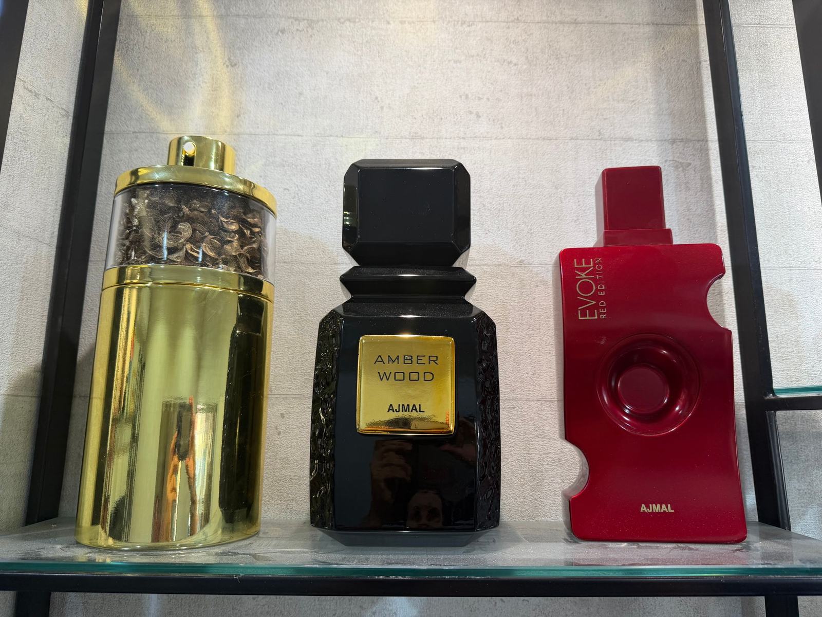 ثلاثة مجسمات عملاقة لزجاجات عطور مصنوعة بالطباعة ثلاثية الأبعاد بالراتنج لشركة Ajmal Perfumes: زجاجة أسطوانية ذهبية، وزجاجة Amber Wood السوداء، وزجاجة Evoke Red Edition الحمراء، على رف زجاجي