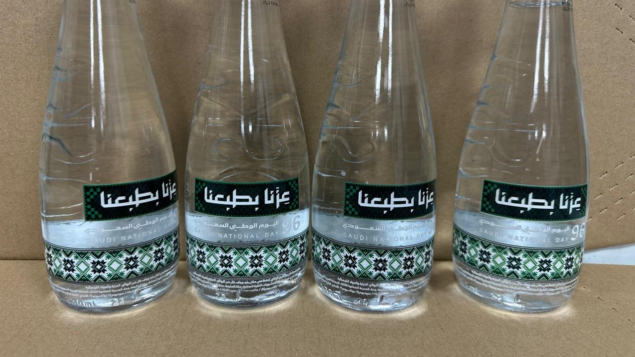 Four filled clear glass water bottles printed with an Arabic national day design, packed together in a carton