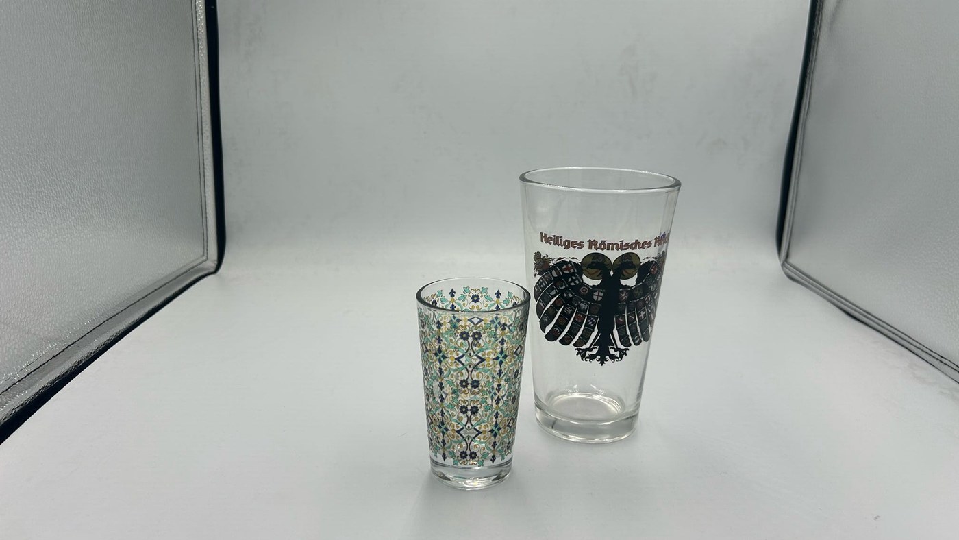 Two printed drinking glasses, a small patterned tumbler and a taller glass carrying a black crest