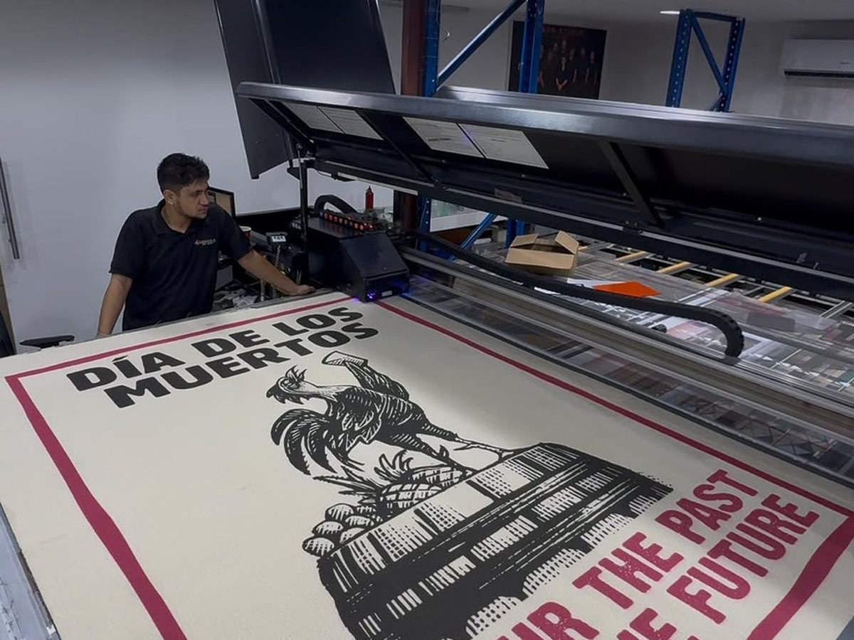 السجادة الثانية على الماكينة، مطبوعة بديك أسود بأسلوب الحفر فوق أفق مدينة وإطار قرمزي، مع عبارة Día de los Muertos