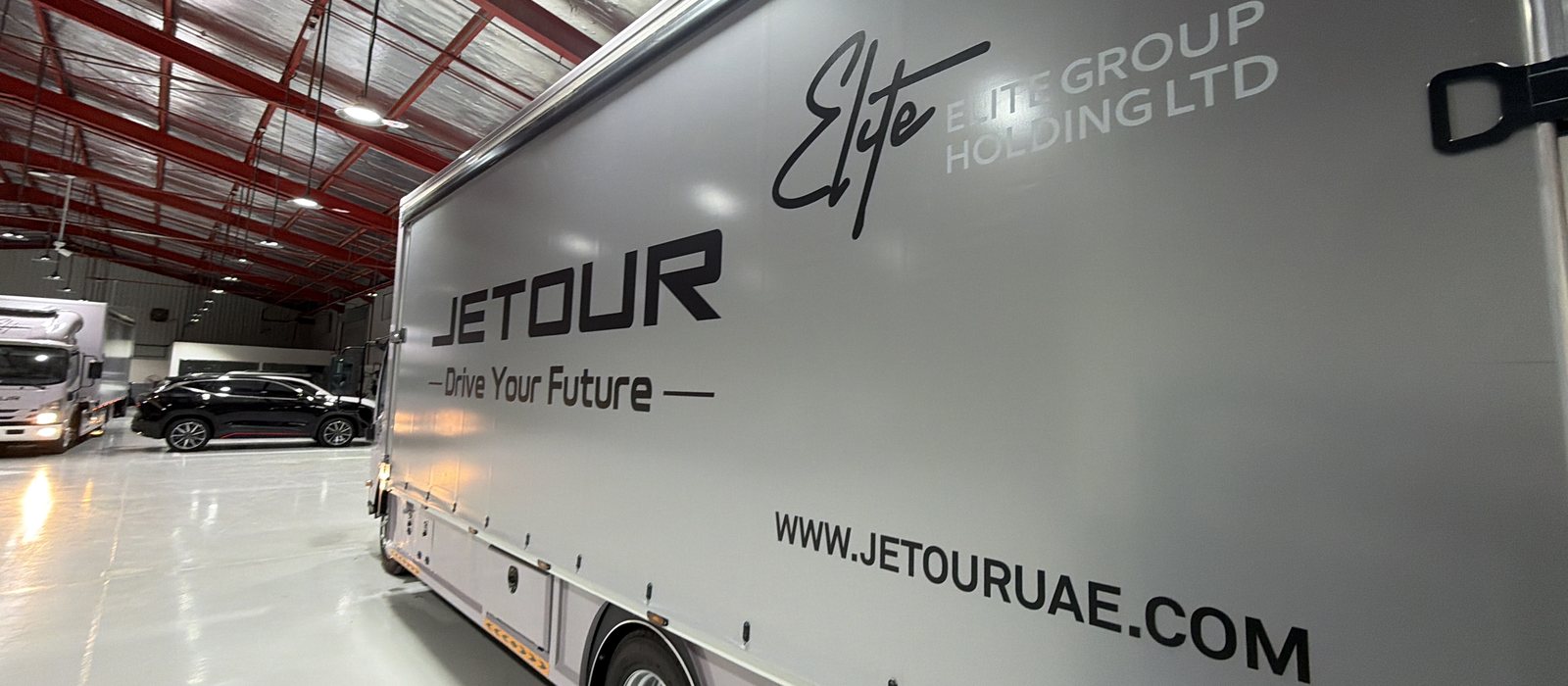 مشمّع جانبي رمادي لشاحنة JETOUR مطبوع بكلمة JETOUR وعبارة Drive Your Future وشعار Elite Group Holding بالأسود والأبيض، وعنوان www.jetouruae.com