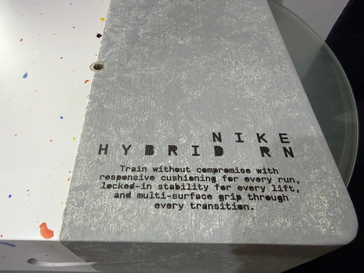 لقطة قريبة للرخام المطبوع: كلمة NIKE وعبارة HYBRID RN بحروف كبيرة متباعدة فوق النص الترويجي، وملمس الحجر يظهر من خلال الحبر الأسود