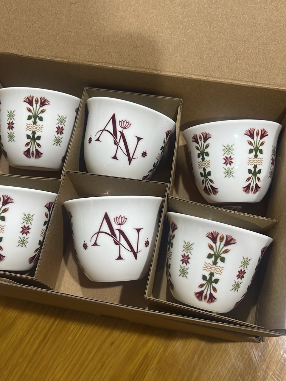 فناجين قهوة عربية من البورسلين في علبة هدايا كرتونية مقسّمة: اثنان مطبوع عليهما حرفا AN باللون العنابي مع زهرة لوتس صغيرة، والبقية بسيقان زهور طويلة حمراء وخضراء وخطوط ذهبية متموجة ونقوش نجوم مطرّزة