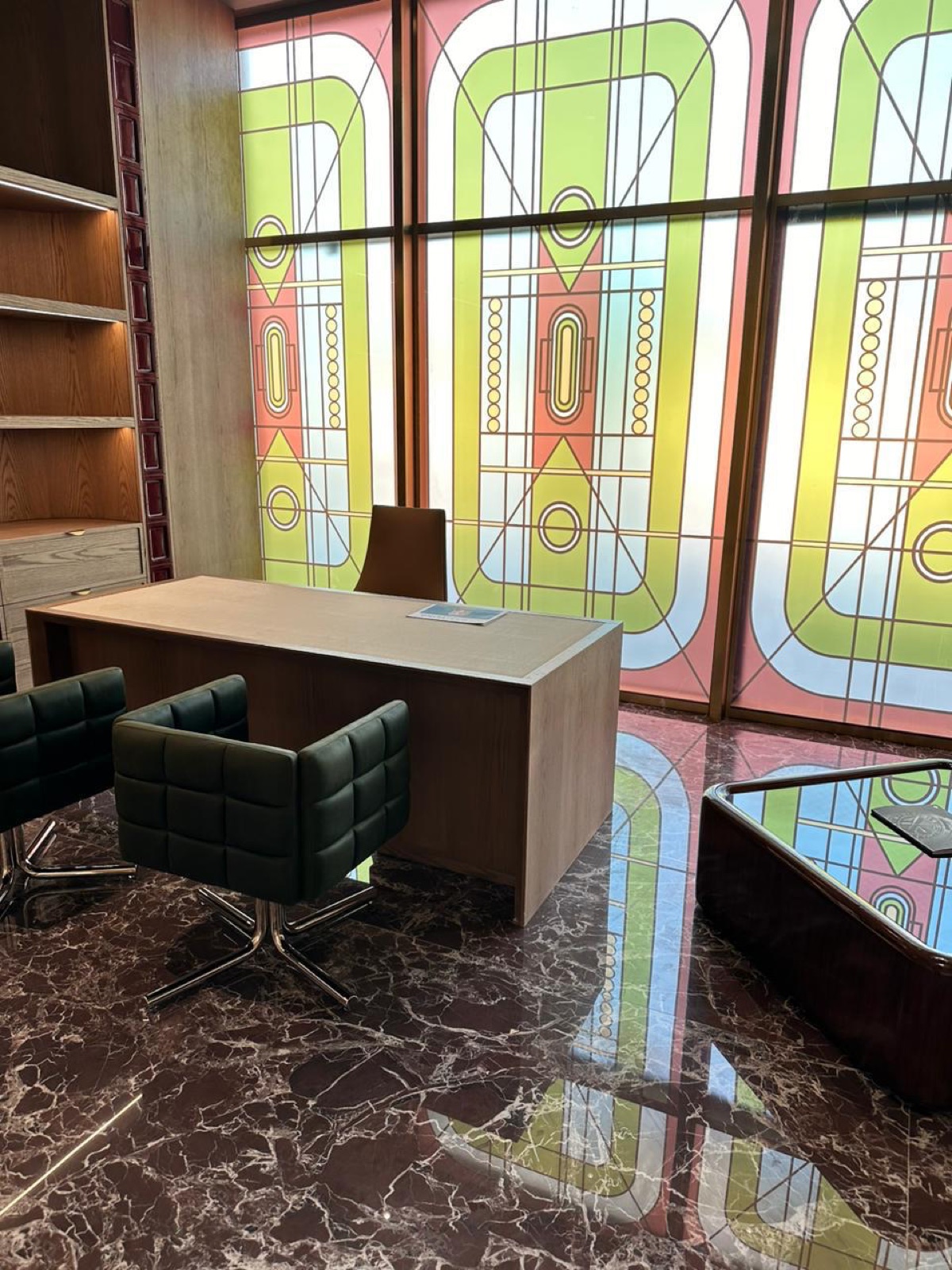 A floor-to-ceiling backlit glass wall printed with pastel Art Deco motifs in a furnished office