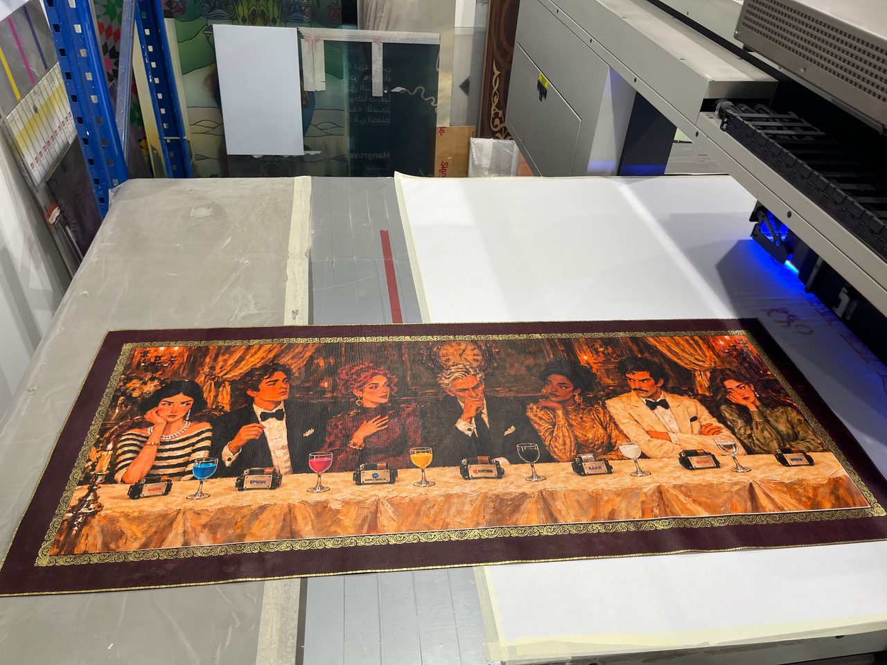 A natural leather panel printed in full colour with a dinner-party painting inside a gold-coloured border, lying on a UV flatbed printer with the lamp lit