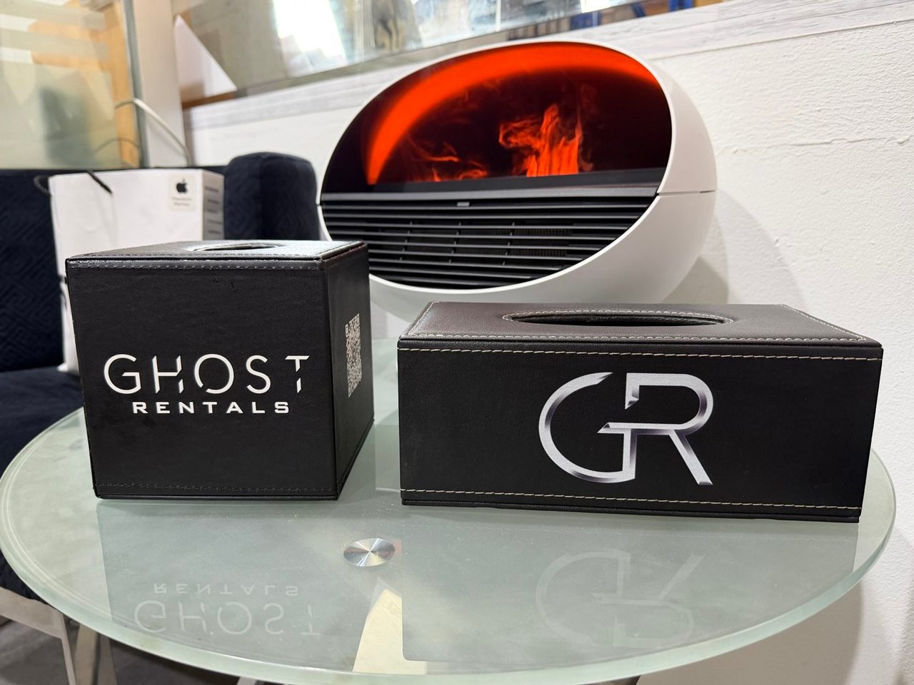 Two black PU leather tissue boxes with contrast stitching on a glass table, the cube printed with a white wordmark and the long box with a monogram in a grey-to-white gradient that reads like polished metal