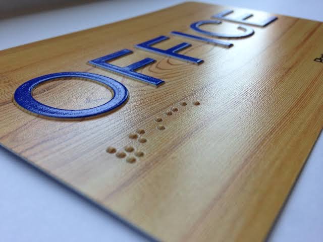 A wood plaque printed with raised metallic blue lettering and a row of tactile dots