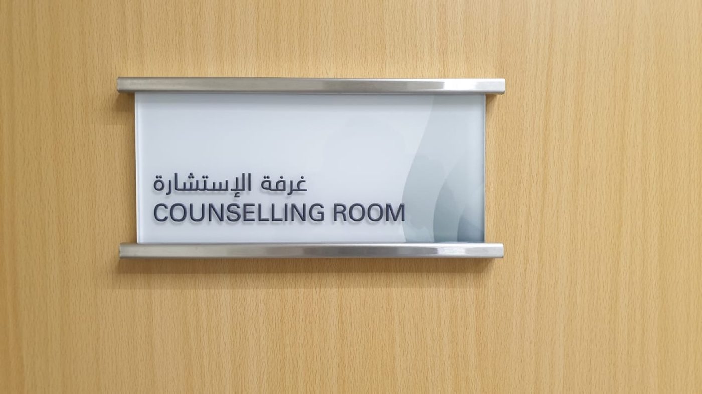 Bilingual Counselling Room door nameplate, dark lettering on a white panel with a soft grey wave, held between brushed-metal rails on a wooden door