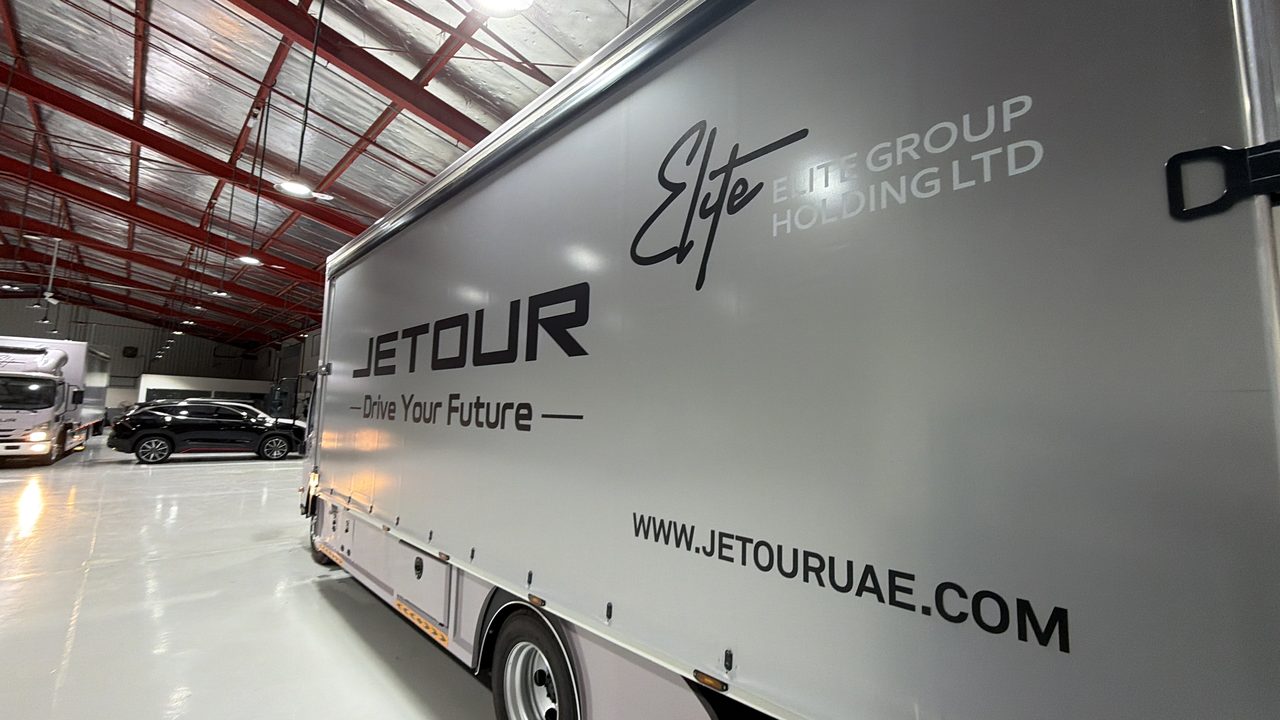 The grey tarpaulin side of a JETOUR truck printed in black and white with the JETOUR wordmark, Drive Your Future, the Elite Group Holding logo and www.jetouruae.com, parked in a service hall
