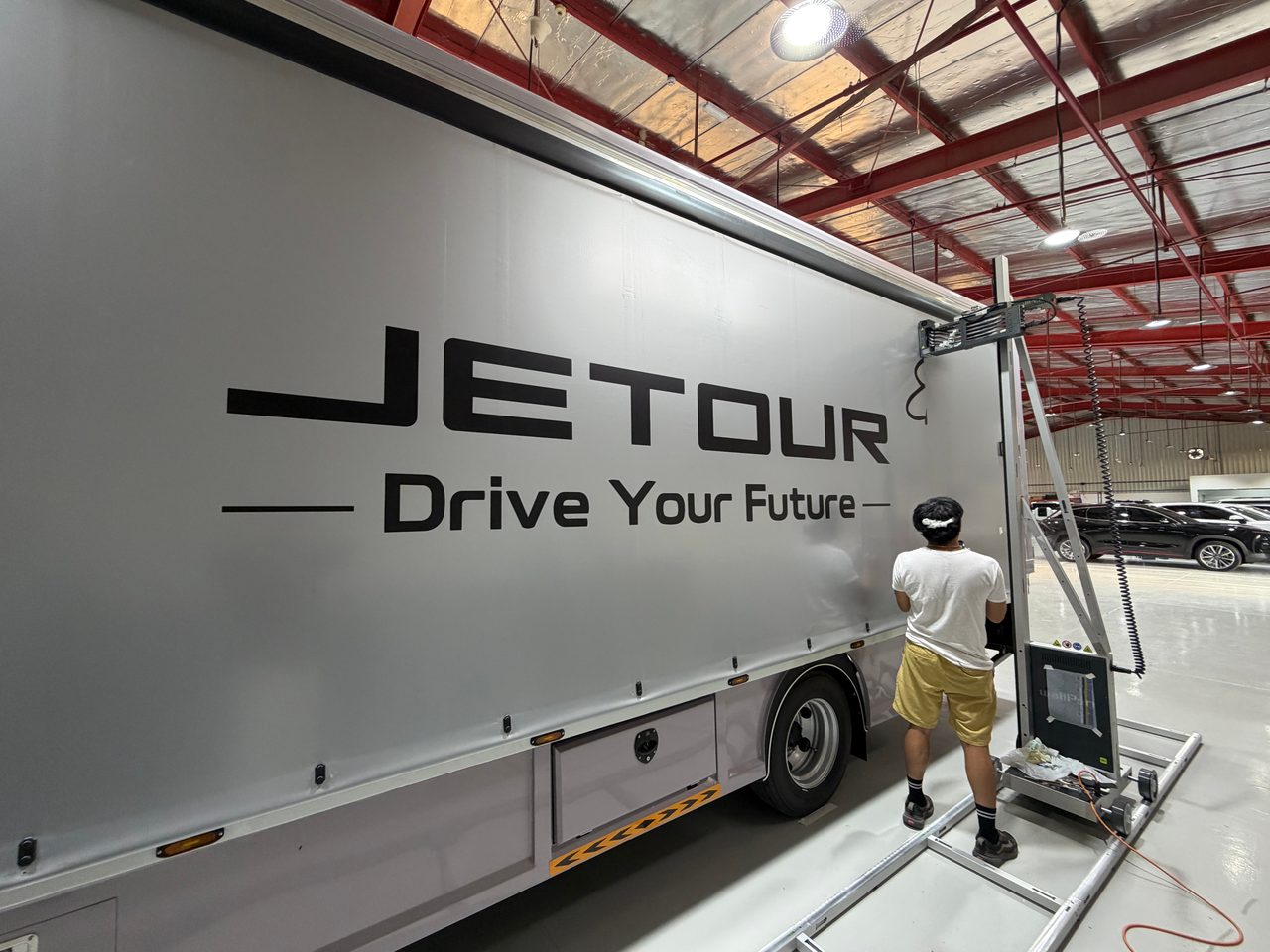A wallPen printer on floor rails at the end of a truck’s grey tarpaulin side, newly printed with JETOUR and Drive Your Future, an operator beside it