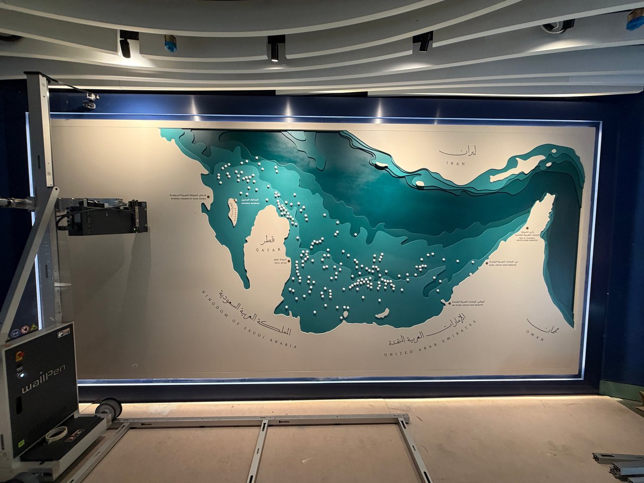 طابعة wallPen بجانب لوح مركّب في متحف يحمل خريطة متعددة الطبقات للخليج العربي تبرز منها كرات تشبه اللآلئ، وأسماء الدول والمدن مطبوعة بالعربية والإنجليزية