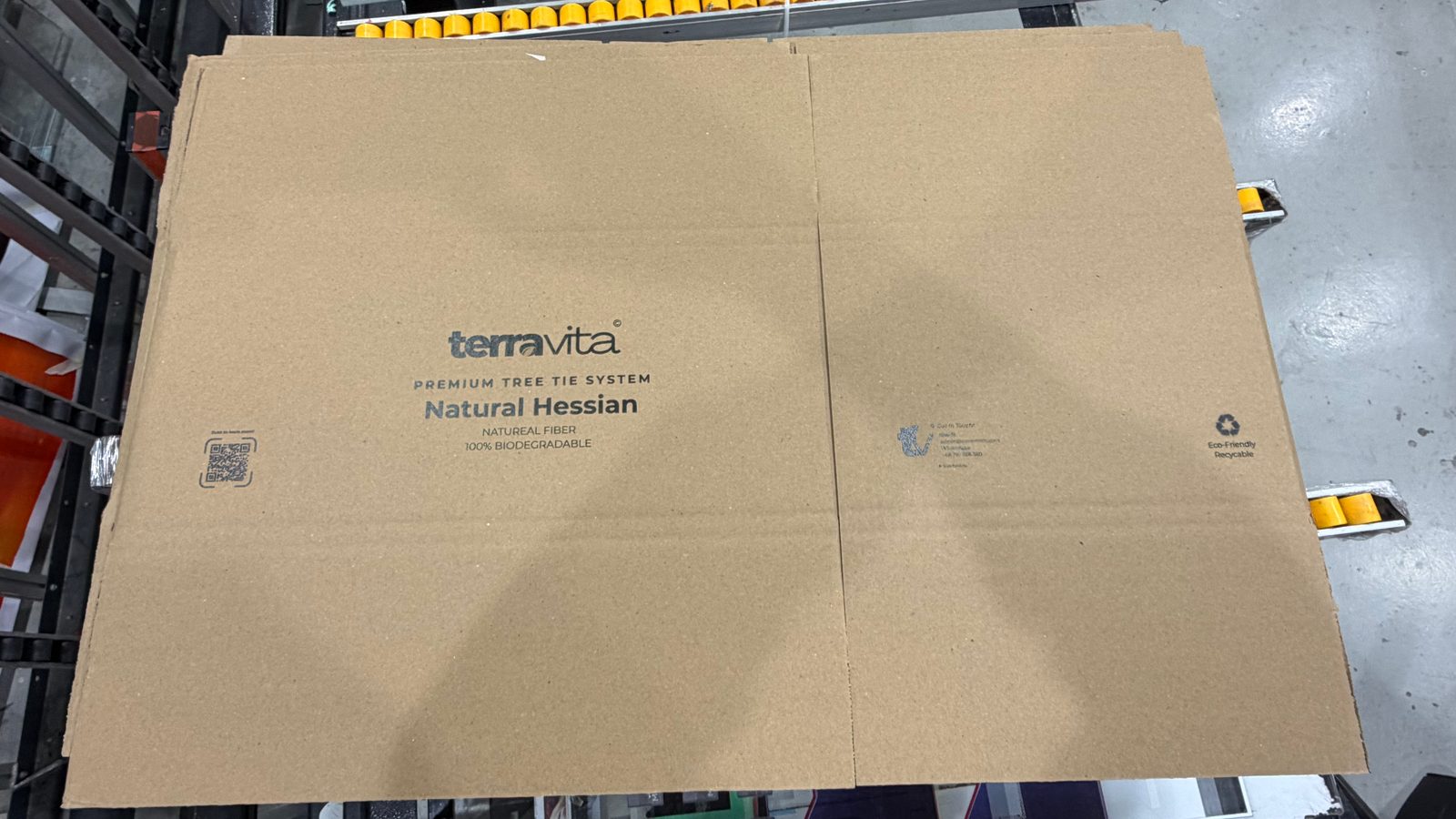 A brown corrugated carton lying folded flat, printed in one dark ink with a brand logo, product name and QR code on one panel and contact details and a recycling mark on the next