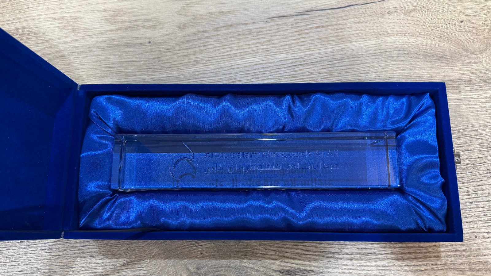The same blue velvet box open, lined with blue satin, holding a clear crystal desk name plate whose name in Arabic and English is engraved rather than printed