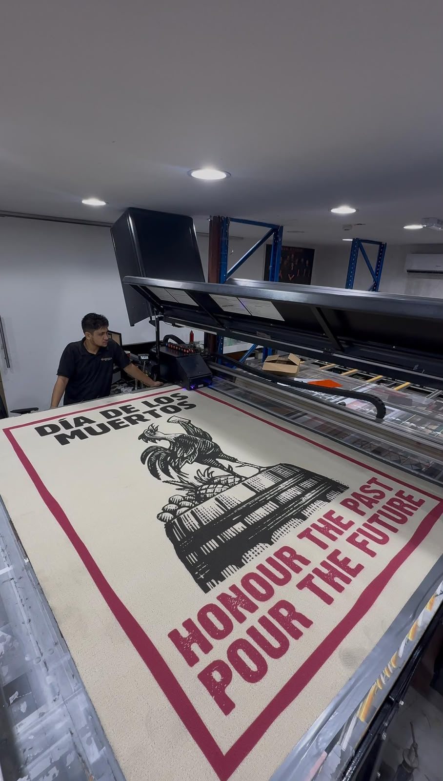A cream pile rug on the flatbed printed with a black linocut rooster above a city skyline and a crimson border, with the operator at the machine behind it