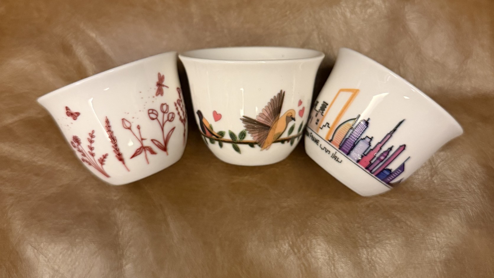 Three printed porcelain qahwa cups, one with a Dubai skyline, one with birds on a branch and one with a botanical pattern