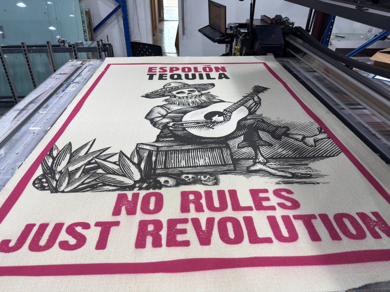 An Espolòn Tequila rug on a UV flatbed: a black linocut skeleton mariachi, agave leaves and skulls inside a magenta border on cream pile