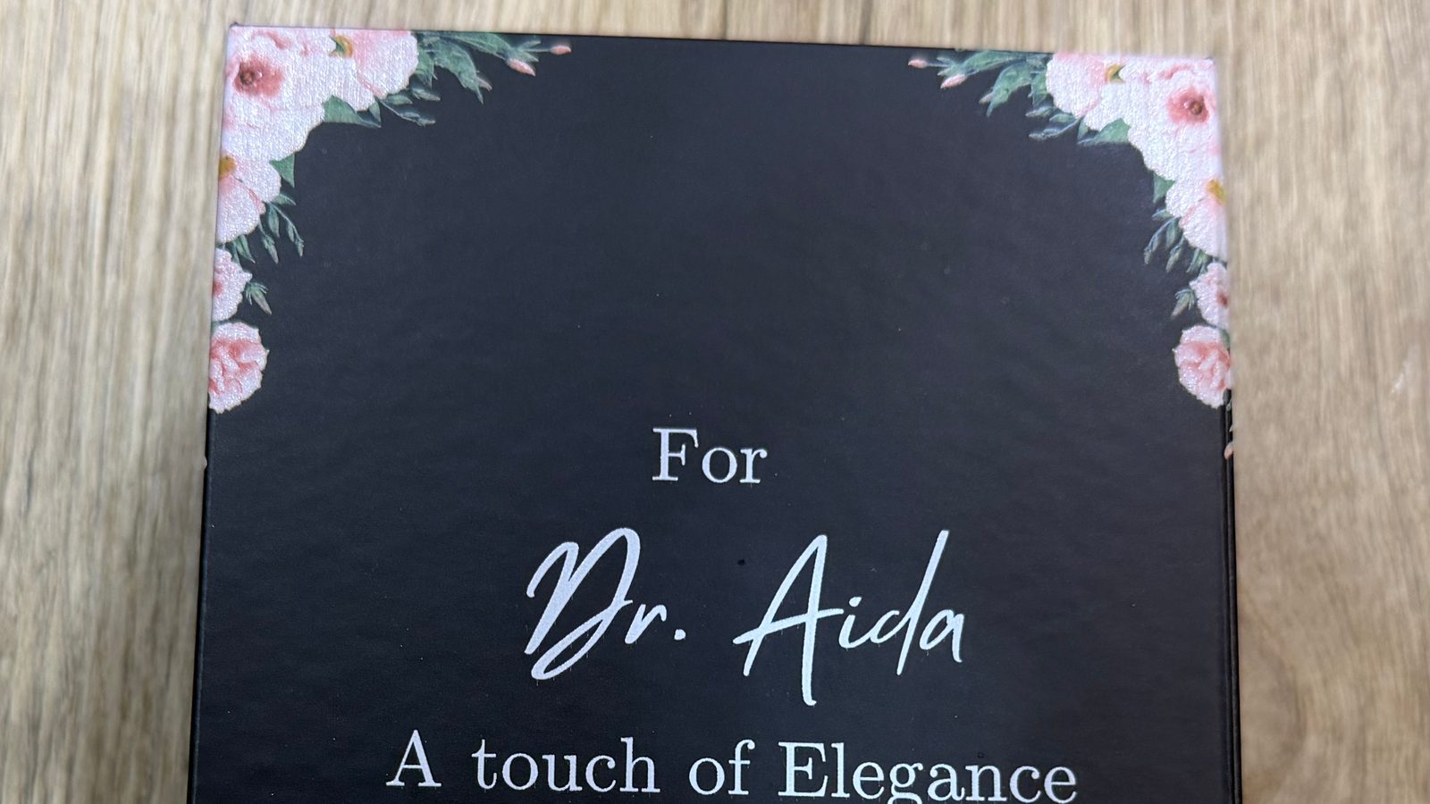 Close-up of a black gift box lid with a recipient's name in white script and a message in white lettering, framed by pink flowers in the two top corners