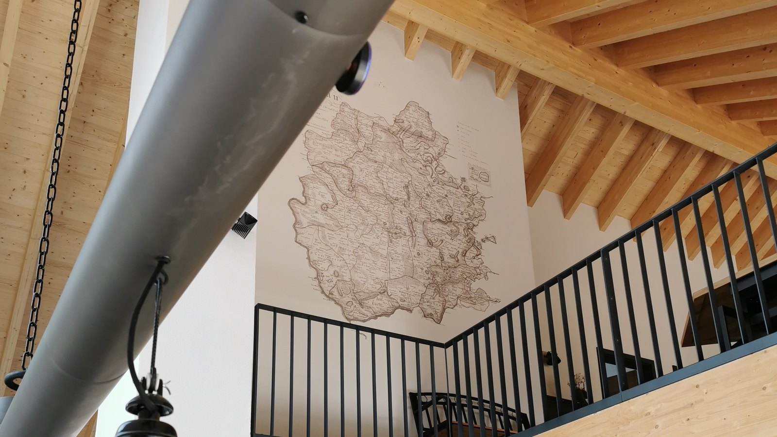 An antique-style map printed directly onto a tall stairwell wall in a home, seen past the mezzanine rail and exposed timber beams