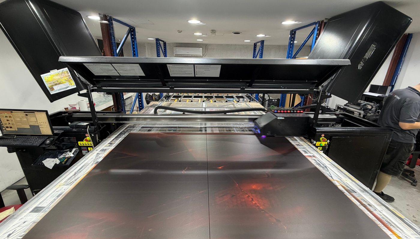 لوحان كبيران من الزجاج على سطح طابعة UV المسطحة IQDEMY EVO 2500 في مستودع صن ستوديو بالقوز، وعربة الطباعة تتوهج بلون بنفسجي على جسرها، وعامل تشغيل إلى اليمين، ورفوف تخزين زرقاء على امتداد الجدران
