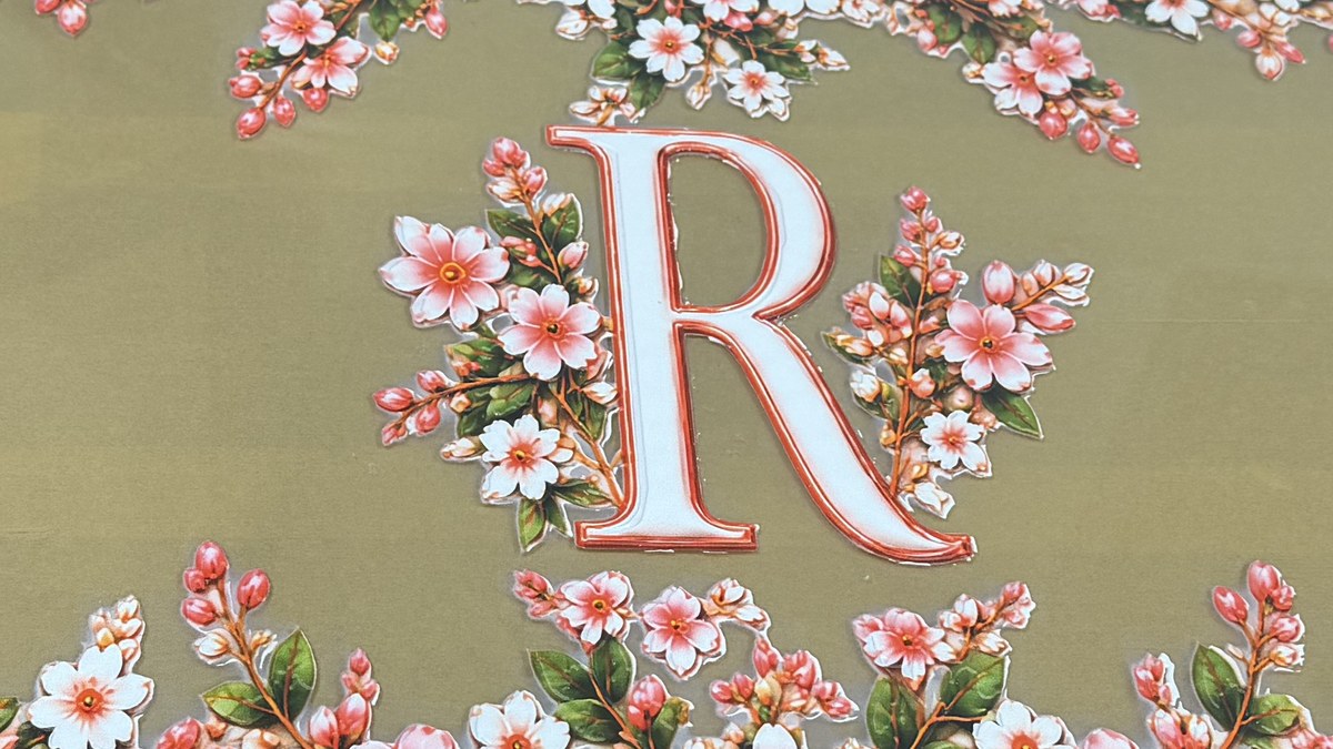 Close-up of a monogram R in white with a red outline, with blossom on either side, on gold acrylic