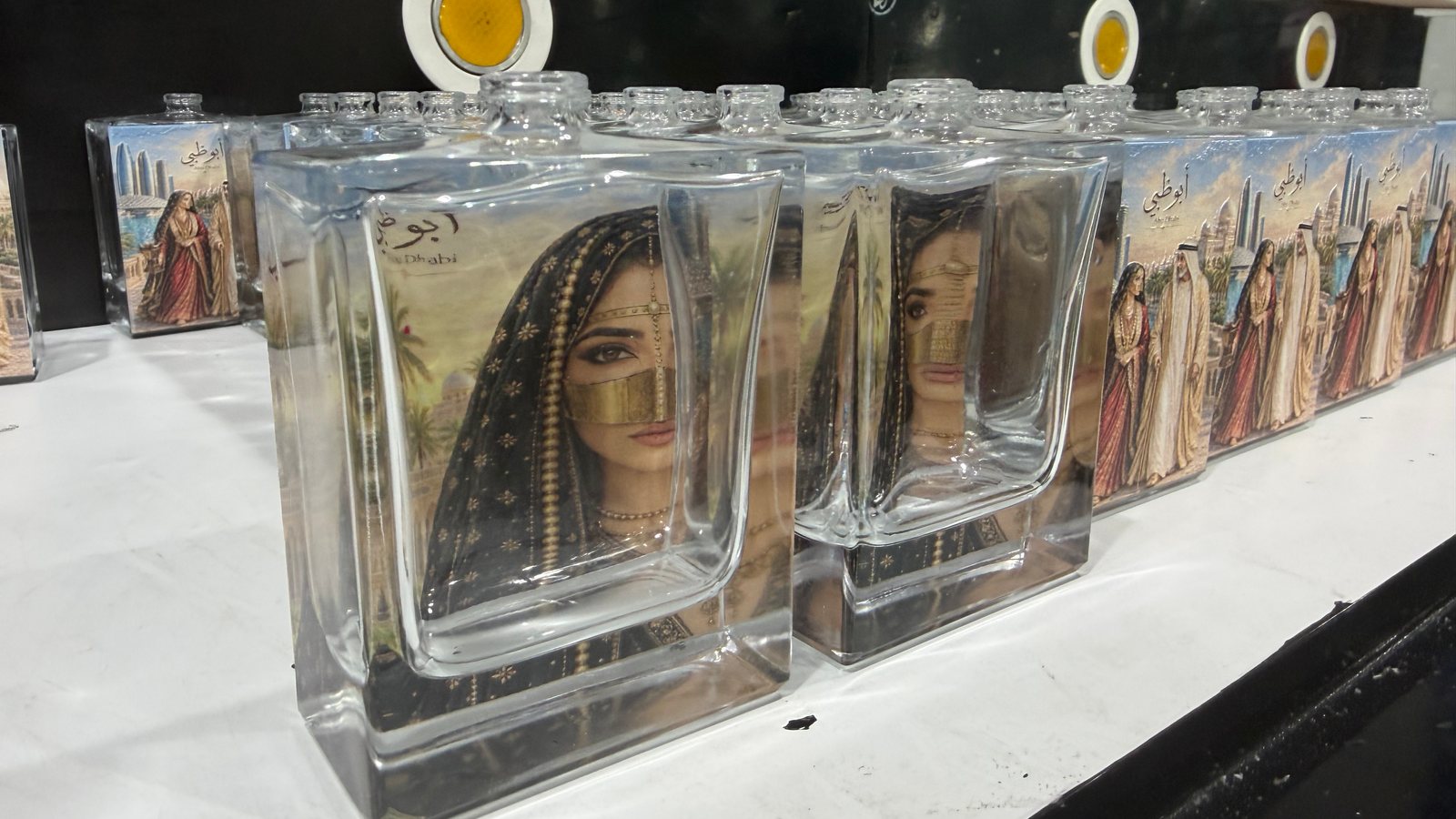 زجاجات عطور مسطّحة مطبوعة مصوّرة بزاوية مائلة، والتصميم يظهر عبر الزجاج الشفاف من الوجه غير المطبوع، والزجاجة الأمامية تحمل صورة لامرأة ترتدي البرقع الذهبي بدل الثنائي