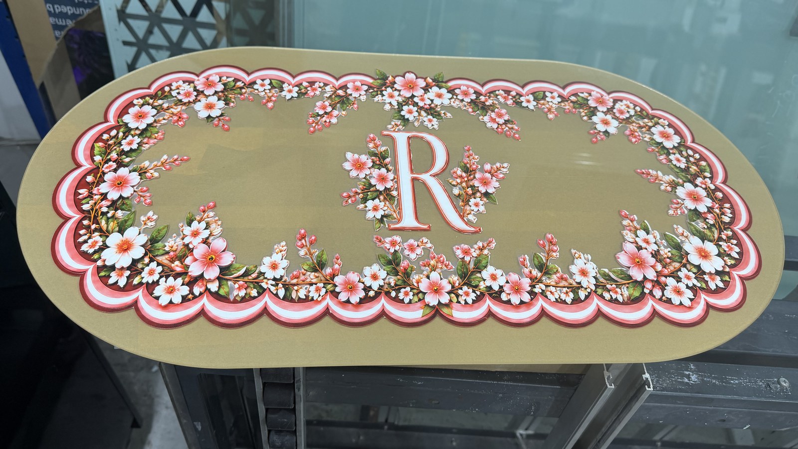 An oval piece of gold acrylic printed with a wreath of pink and white cherry blossom, a scalloped border and a monogram R at its centre, in the Sun Studio workshop in Dubai