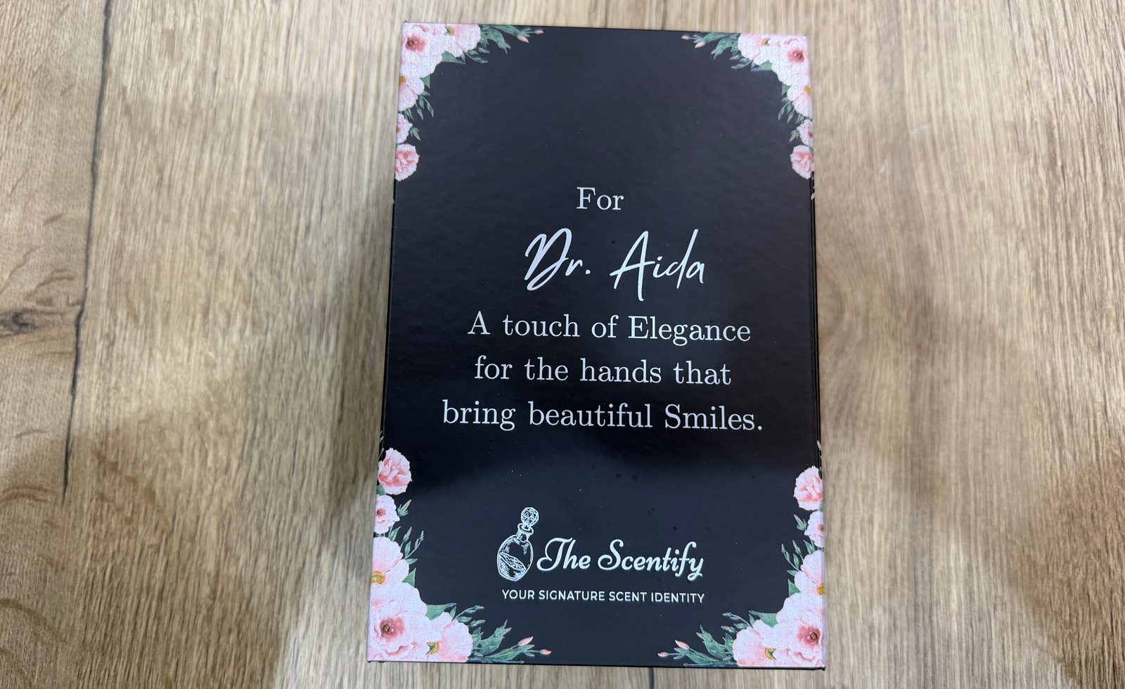 Black gift box lid printed with pink flowers in each corner, a personalised message in white lettering and a perfume brand logo, on a wooden table