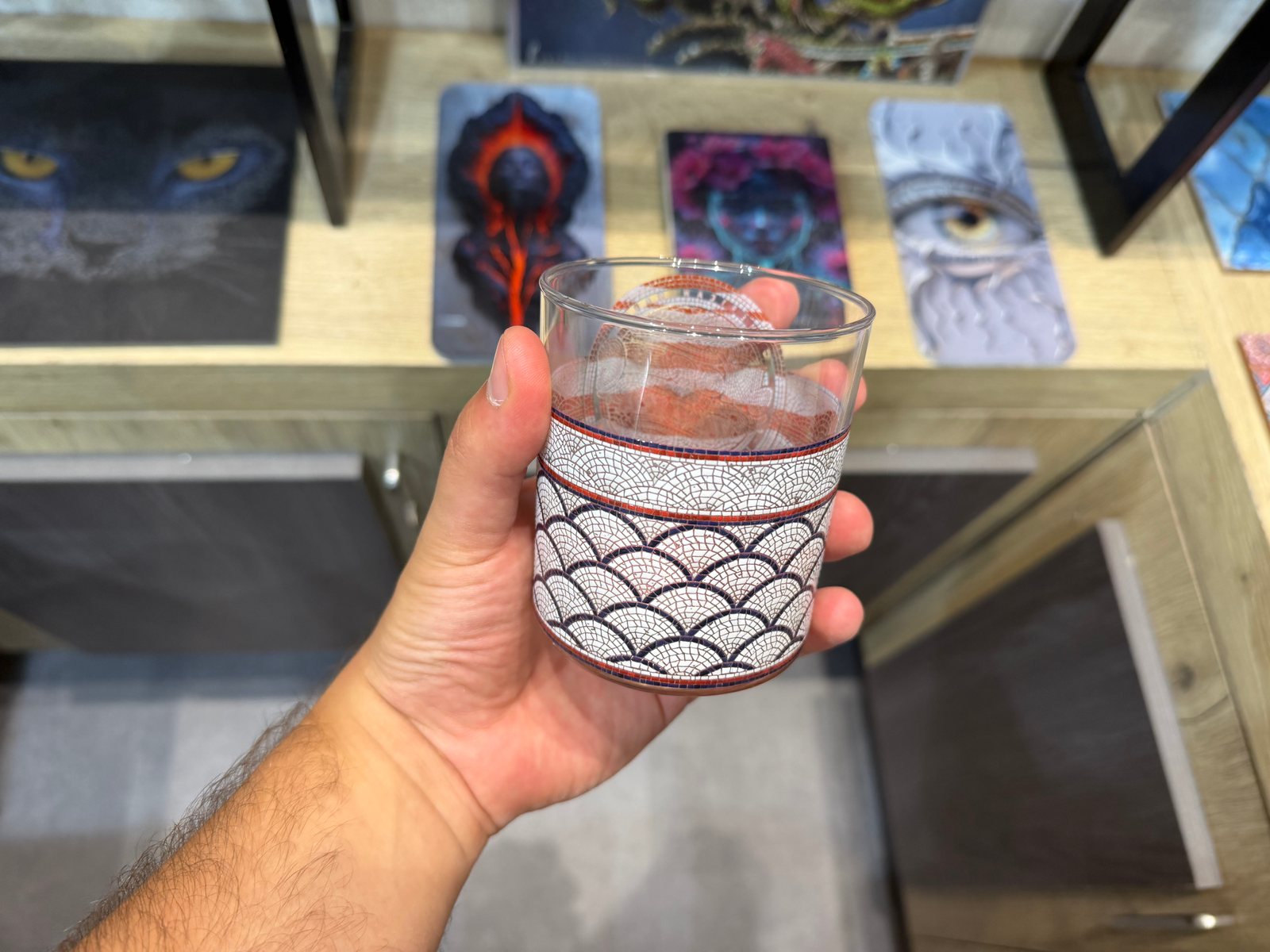 A clear glass tumbler held in the hand, printed with a mosaic-style scalloped scale pattern in white outlined in dark blue, with rust-red mosaic bands edged in navy at the rim