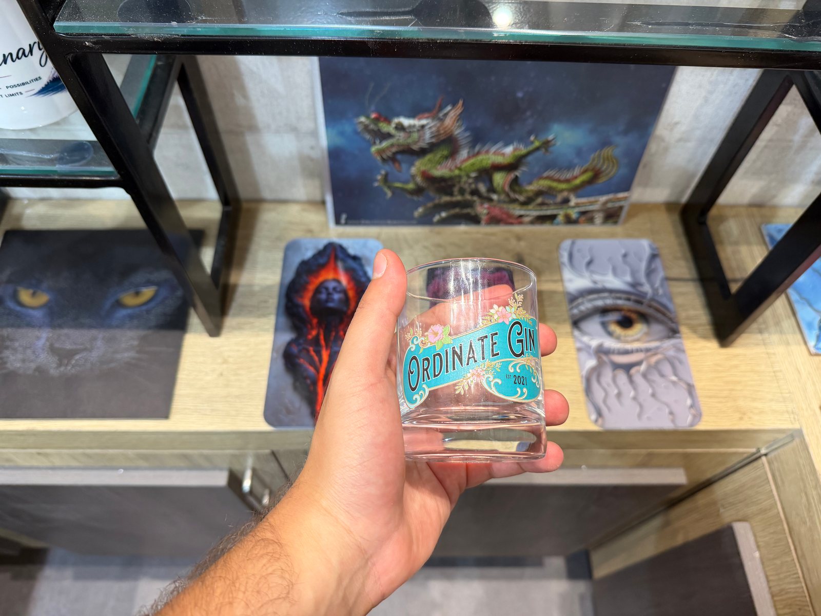 A clear glass tumbler held in the hand, printed with a turquoise banner label and floral ornament
