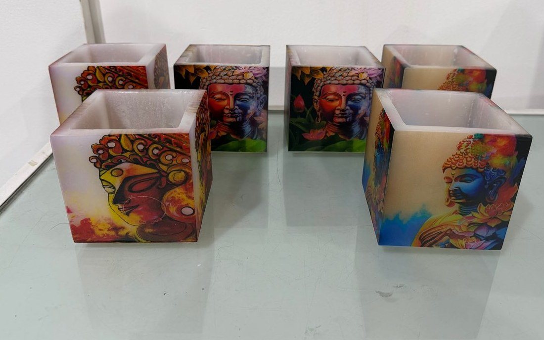 Six square wax candle holders on a glass shelf, printed on their sides with colourful Buddha and portrait artworks