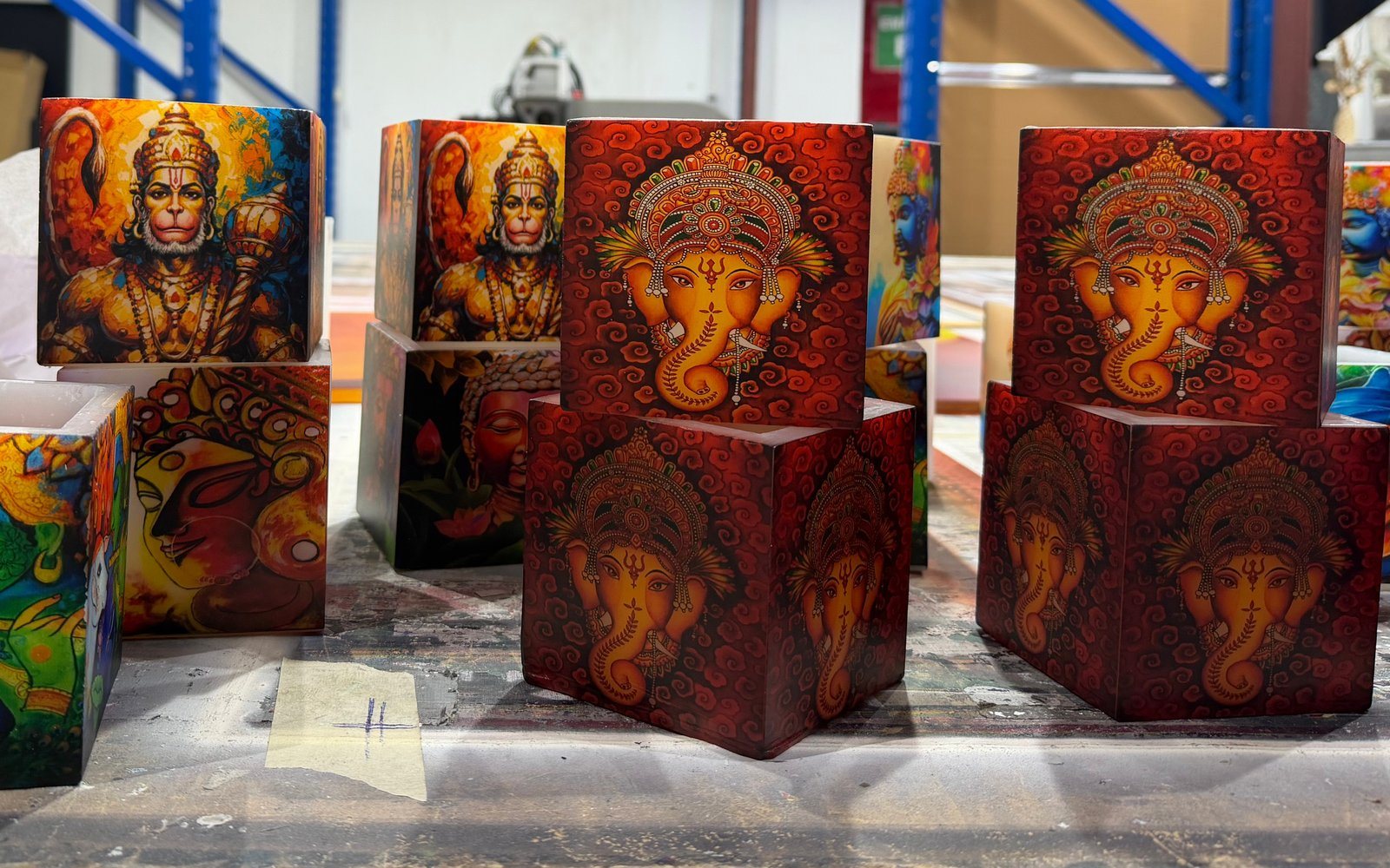 Square wax candle holders stacked on a worktable, printed with Ganesha on red swirls and with Hanuman and Buddha artworks