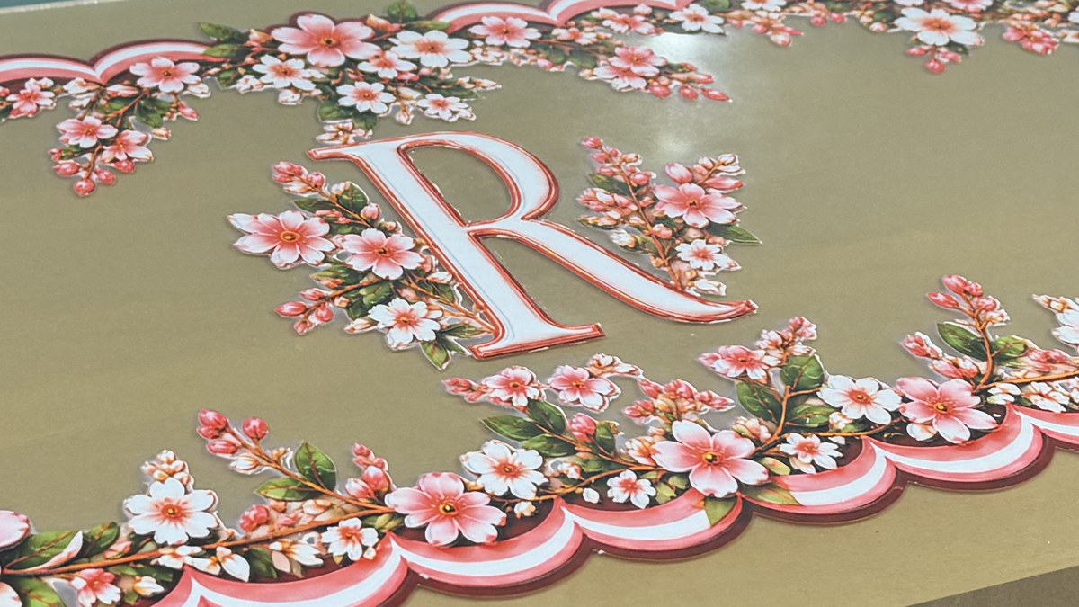 A monogram R in white with a red outline, framed by sprays of blossom, printed on a sheet of gold acrylic