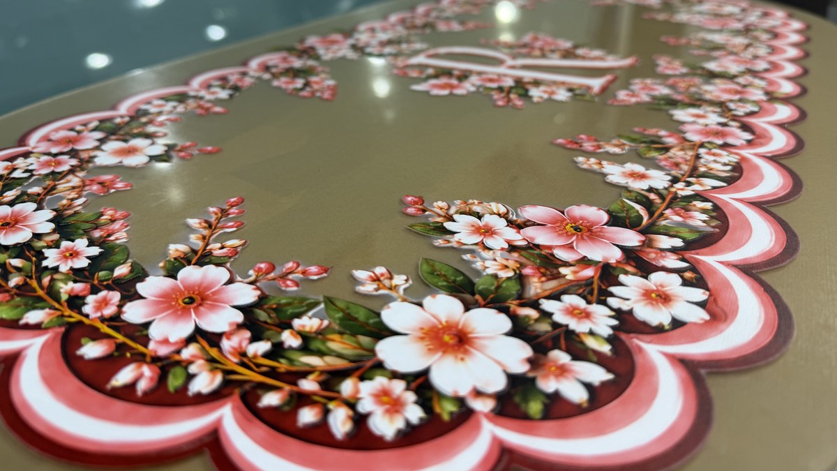 Low-angle close-up of raised pink and white flowers and a scalloped border printed on gold acrylic, with a monogram R out of focus behind them