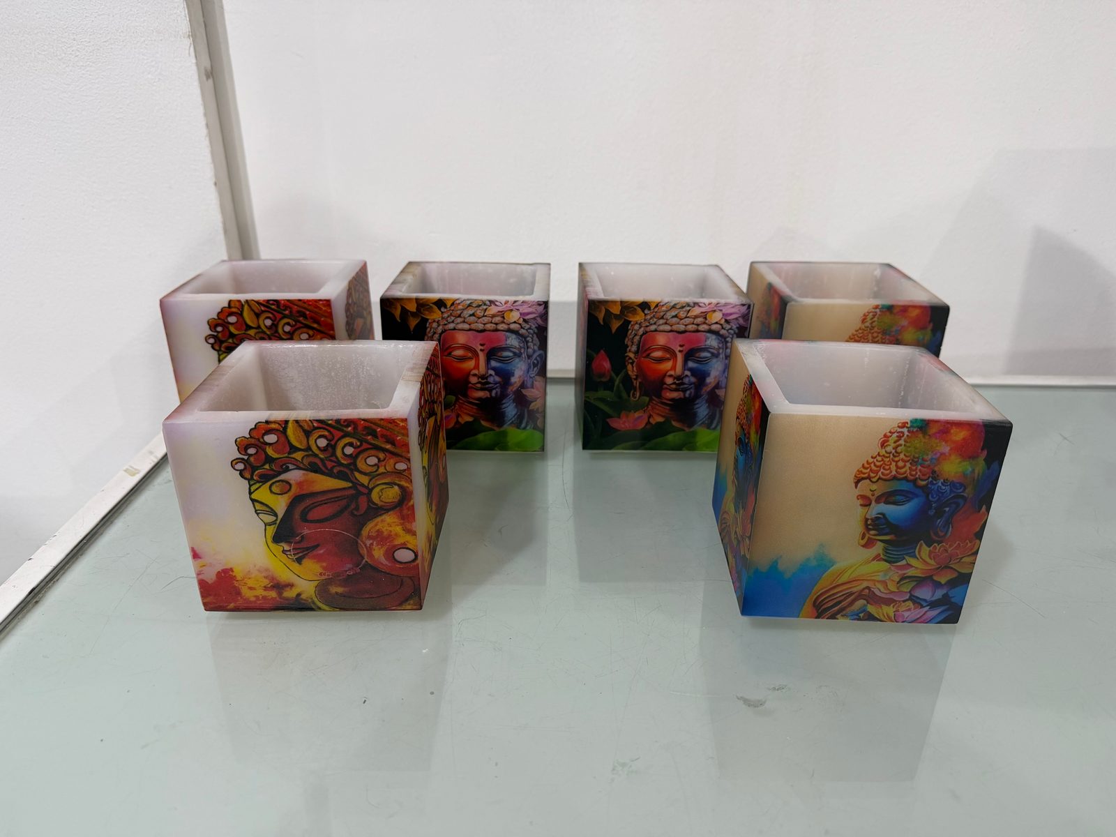 Six square wax candle holders printed with colourful Buddha and portrait artwork, set out on a glass shelf
