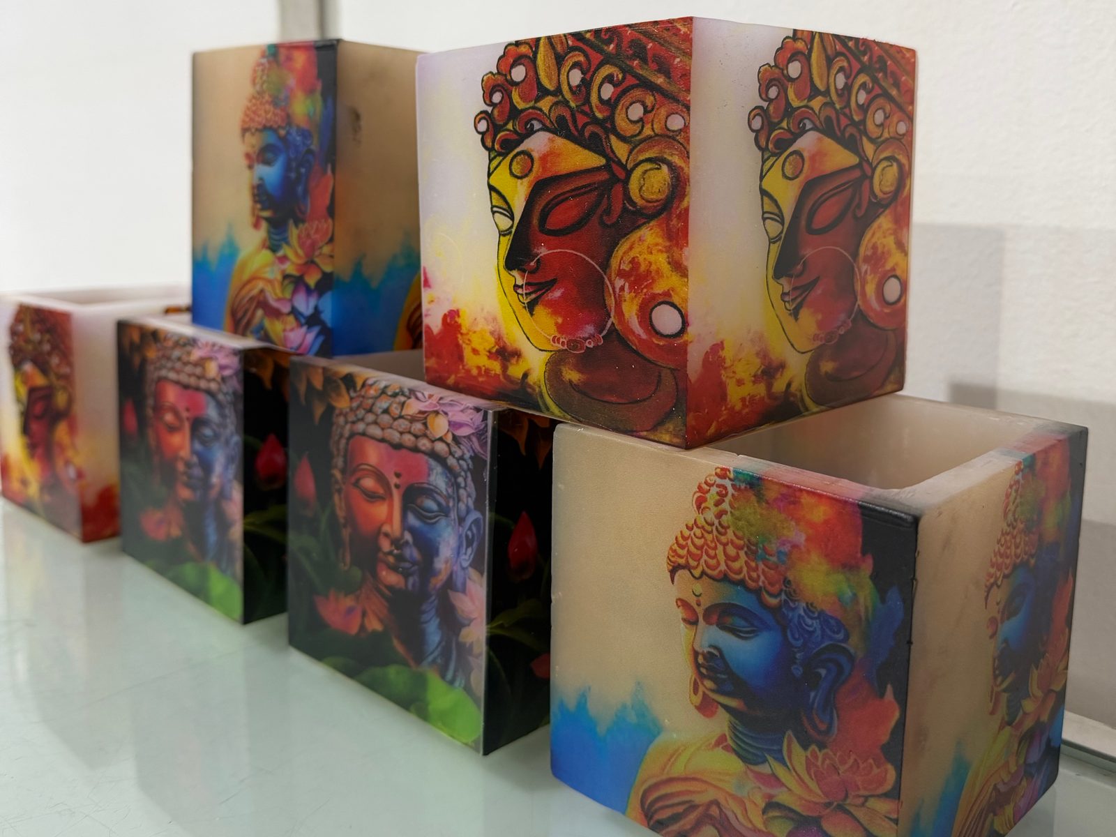 Square wax candle holders printed on their sides with Buddha and portrait artwork, stacked on a glass shelf