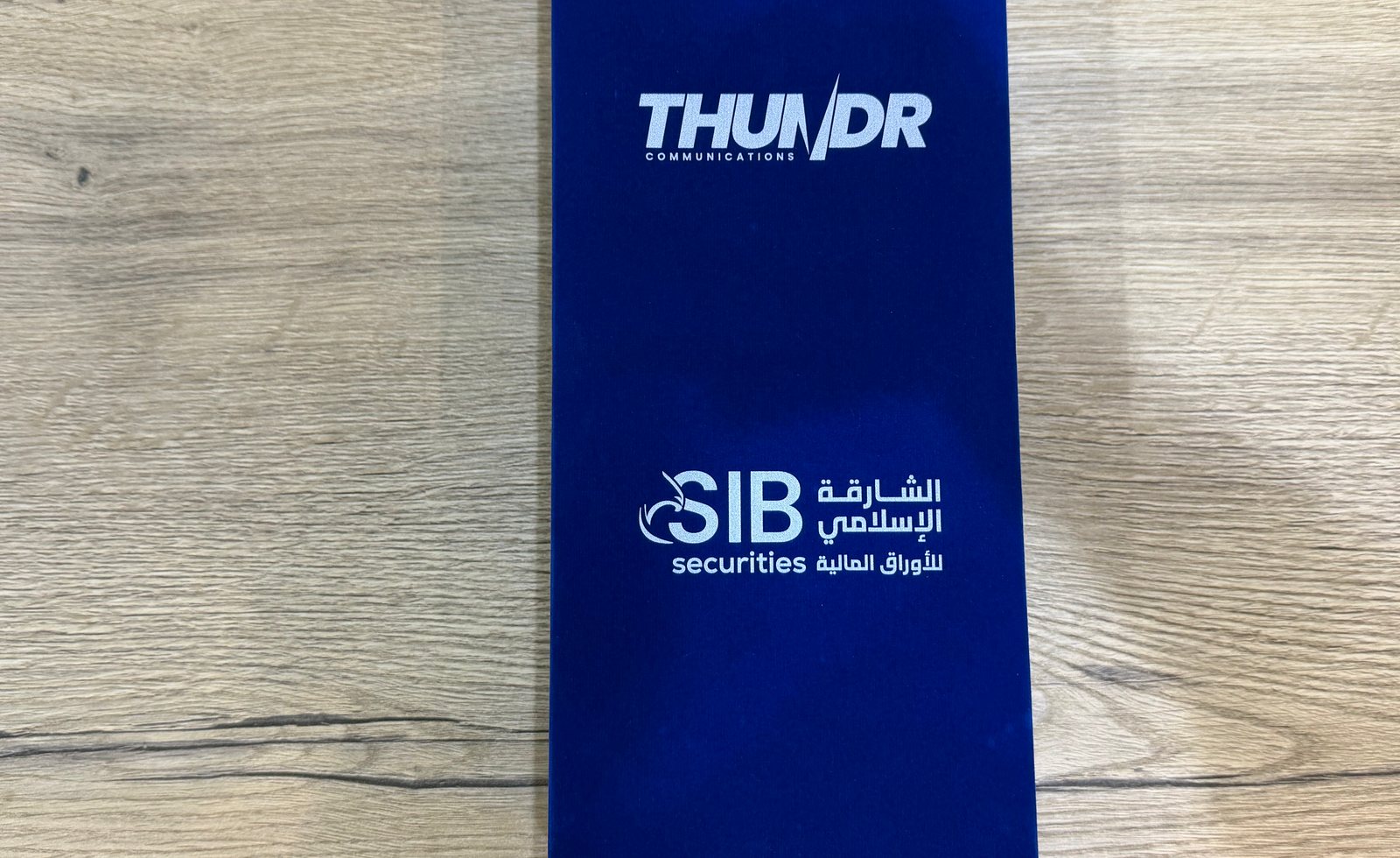 A blue velvet presentation box photographed from above on a wooden surface, two brand logos printed on the lid in white, the lower one set in Arabic and English