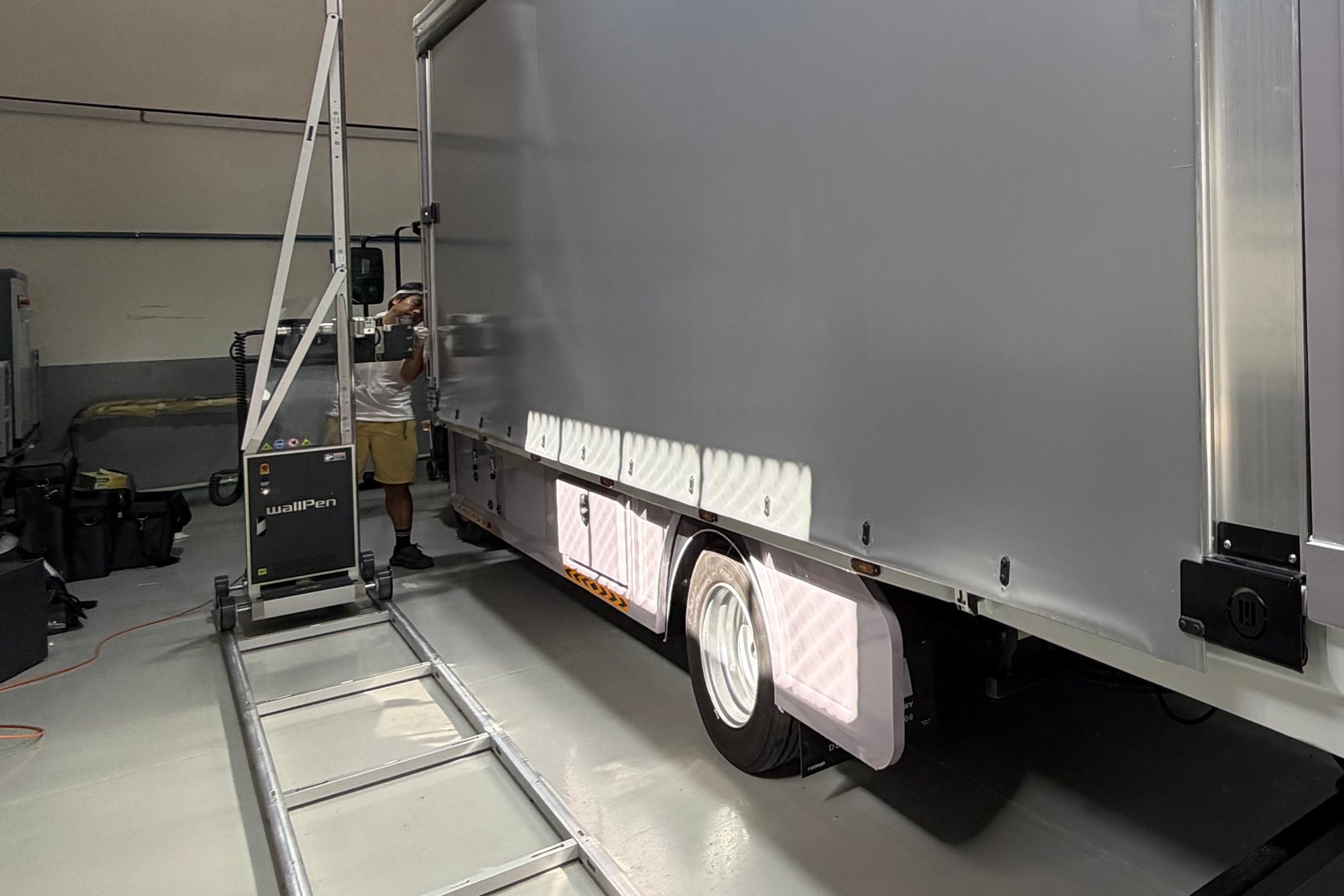 The blank grey tarpaulin side of a truck before printing, wallPen floor rails laid along it and the printer at the far end, an operator beside it