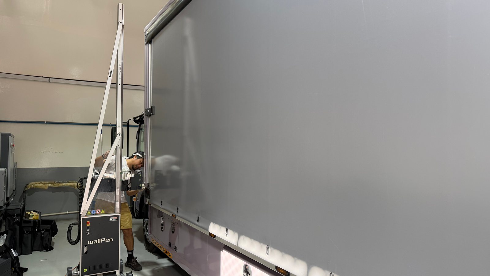 An operator at a wallPen printer standing on rails close to the grey tarpaulin side of a truck, the printed word Drive just visible behind the machine